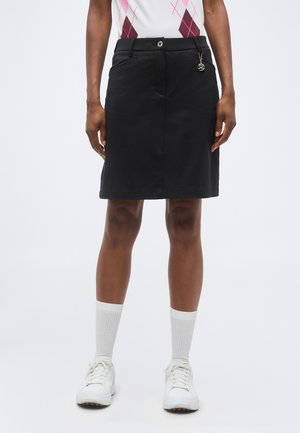 Henkilöllä on musta polvipituinen hame, jossa on etutaskut, nappi ja koristeellinen yksityiskohta, valkoiset sukat, valkoiset tennarit ja kuvioitu paita.