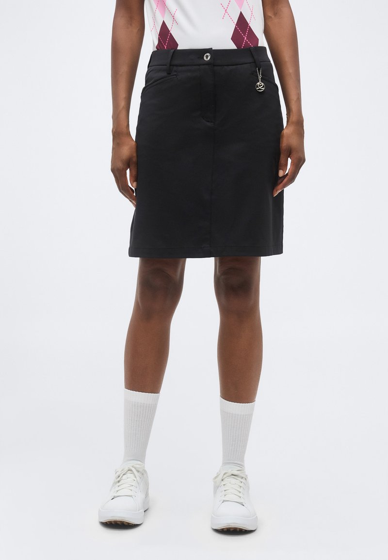 Άτομο με μαύρη φούστα μήκους έως το γόνατο με μπροστινές τσέπες, κουμπί και διακοσμητικό στοιχείο, λευκές κάλτσες, λευκά αθλητικά παπούτσια και μπλούζα με σχέδιο.