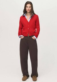 Červený cardigan s knoflíky, vrstvený přes bledě modrou košili, spárovaný s tmavě hnědými širokými kalhotami a hnědými botami.