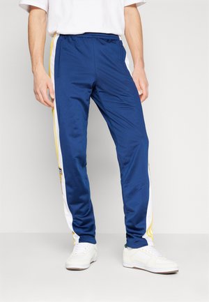 Pantalons de survêtement bleu marine avec des rayures blanches sur les côtés et des accents jaunes. Fabriqués en tissu lisse ; présentent une taille élastique et des poches zippées.