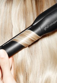 Des cheveux blonds enroulés autour d'un fer à friser sans fil ghd noir, tenu par une main.