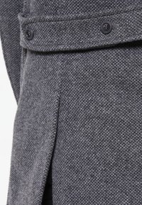 Abrigo de lana gris con una tela texturizada, que presenta un cuello con muesca, dos botones y una abertura lateral para mayor comodidad al moverse.