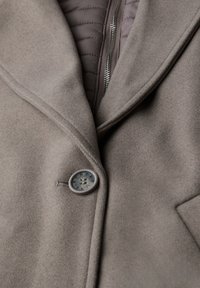 Cappotto grigio con una texture spazzolata, dotato di colletto a risvolto, fodera interna trapuntata e un bottone rotondo con testo, posizionato sul davanti.
