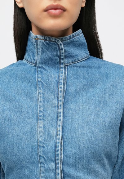 Heleroheline teksajakk kõrge krae, nähtavate õmblustega ja siledapinnaline tekstuur. Ees kinnitusplokk kinnitamiseks.