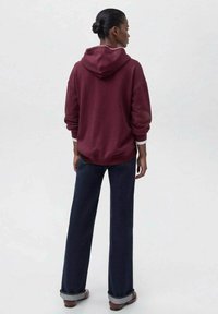 Mujer de pie de espaldas, vestida con una sudadera burdeos, jeans oscuros ajustados y zapatos marrones, contra un fondo claro y liso.