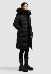 Sort pufferjakke med en faux fur hætte, quiltet design, lynlås foran og sidelommer, kombineret med sorte snørebåndsstøvler og leggings.