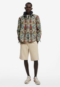 Bunte, mit Blumenmustern versehene Jacke aus leichtem Stoff, beigefarbene Shorts und beigefarbene Sneaker mit grauen Akzenten. Verfügt über eine Kapuze und einen Knopfverschluss.