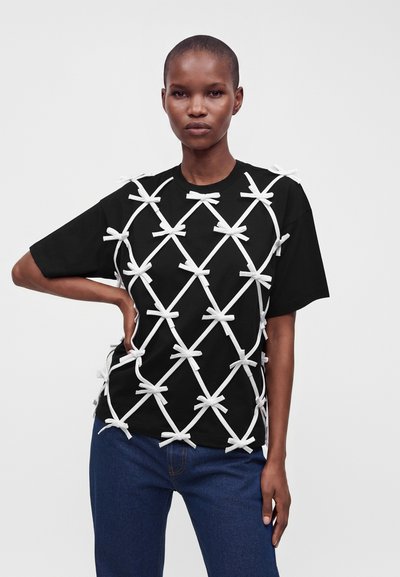 Personne portant un t-shirt noir à manches courtes avec des bretelles blanches croisées nouées en nœuds, associée à un jean bleu, debout avec une main sur la hanche.