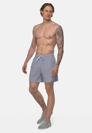 Hemdloser Mann mit Tätowierungen am Arm und Bein, trägt blau-weiß gestreifte Shorts und graue Slip-On-Schuhe, steht vor weißem Hintergrund.
