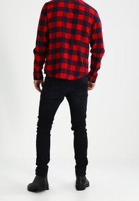 Camisa de franela a cuadros rojos y marinos con puños de botón, combinada con jeans ajustados negros y botines negros, vista desde la parte trasera.
