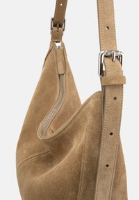 Τσαντάκι ώμου από μπεζ σουέντ με ασημένιο φερμουάρ μερικώς ανοιχτό και ρυθμιζόμενο λουρί με ασημένια αγκράφα σε λευκό φόντο.