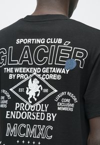 Černé bavlněné tričko s bílými grafikami, včetně nápisu „Sporting Club Glacier“ a lvího emblému. Na rameni je texturovaná modrá nášivka.