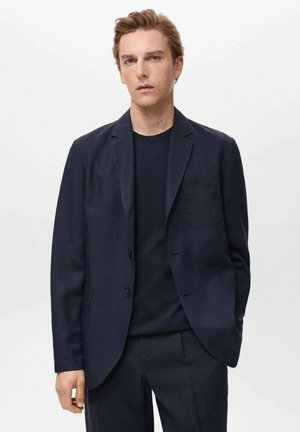 Marineblazer mit einem Einknopfverschluss, Revers und einer Brusttasche. Kombiniert mit einem marineblauen Rundhalsausschnittpullover, taillierter Schnitt. Glatte Textur.