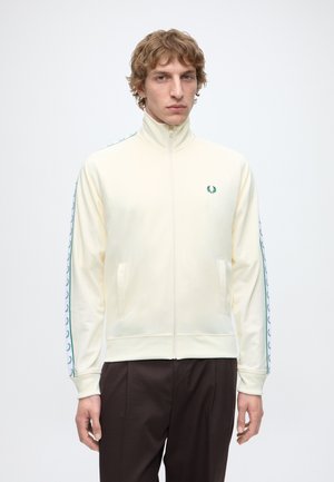 Mann mit lockigem Haar trägt eine cremefarbene Trainingsjacke mit Reißverschluss und grünen Lorbeer-Logos an Ärmeln und Brust, kombiniert mit dunkler Hose.