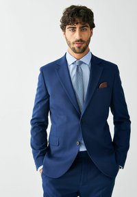 Tengerészkék öltöny strukturált kivitelben; bemetszett hajtókával, egy gombbal és egy mellzsebbel, barna zsebkendővel.