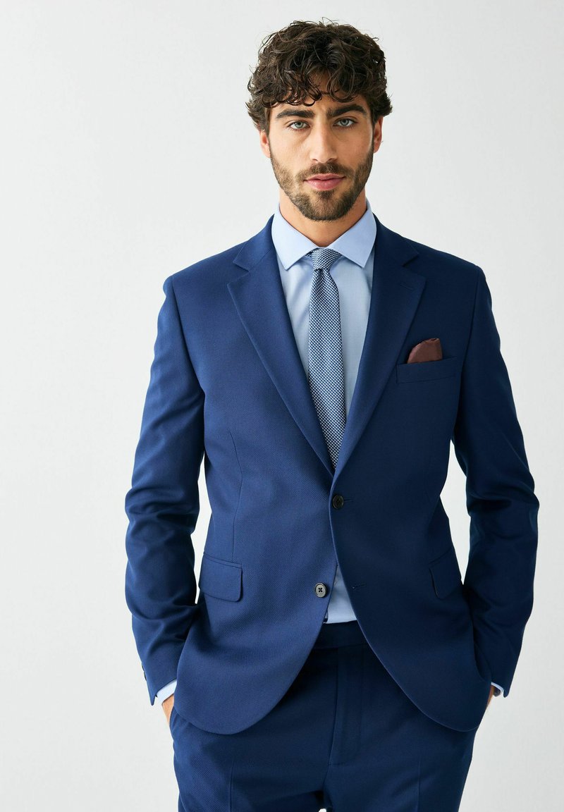 Tengerészkék öltöny strukturált kivitelben; bemetszett hajtókával, egy gombbal és egy mellzsebbel, barna zsebkendővel.