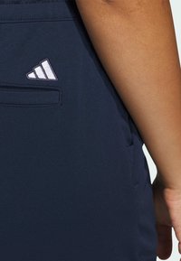 Mörkblå sportshorts i en slät textur, prydda med en liten vit Adidas-logotyp ovanför en sidoficka.