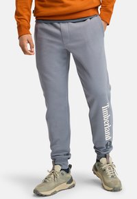 Graue Sweatpants mit Seitentaschen, elastischem Bund, schmal zulaufenden Bündchen und einem weißen "Timberland"-Logo am linken Bein. Glatte Textur.