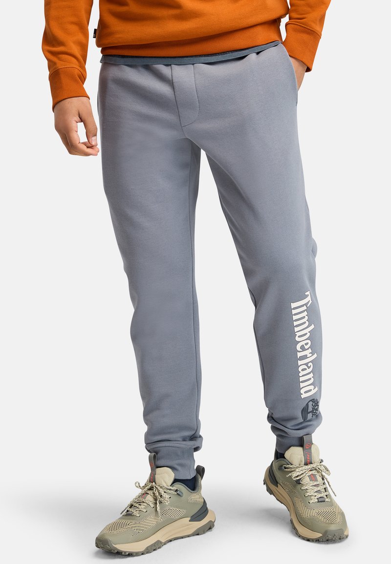 Graue Sweatpants mit Seitentaschen, elastischem Bund, schmal zulaufenden Bündchen und einem weißen "Timberland"-Logo am linken Bein. Glatte Textur.