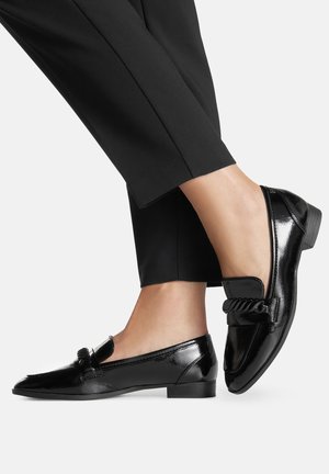 Mocasines negros de cuero brillante con una correa trenzada, usados con pantalones negros recortados, pies cruzados sobre un fondo claro y liso.