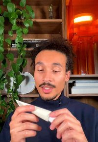 Mann med krøllete hår og bart undersøker et hvitt rør, stående foran treskap med bøker, planter og en oransje lampe.