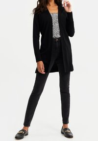 Svart cardigan med ribbade detaljer, bärs över en mönstrad topp och matchas med svarta jeans. Svarta loafers med kedjedetalj fulländar outfiten.