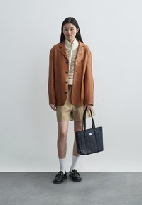 Sacou oversized maro, pulover tricotat crem, pantaloni scurți khaki, geantă tote neagră. Pantofiloaferi negri și șosete albe completează ținuta. Fundal neutru.
