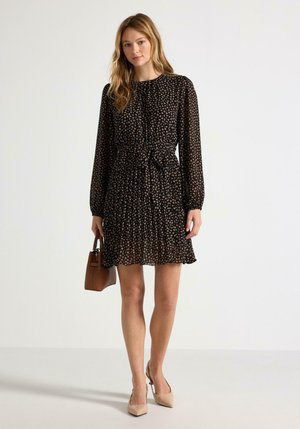 Donna che indossa un vestito lungo a maniche lunghe nero a fiori, tacchi beige e che porta una piccola borsa marrone, in piedi davanti a uno sfondo bianco semplice.