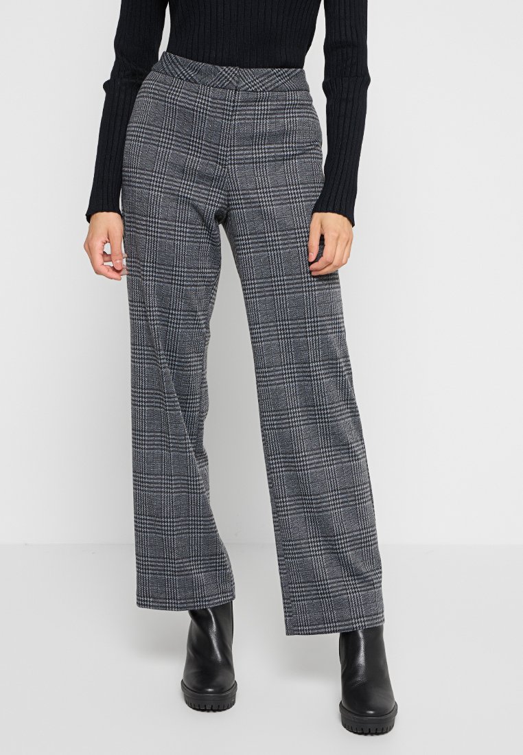 Grau karierte Hose mit hoher Taille, weitem Schnitt und strukturierter Stoffqualität. Kombiniert mit schwarzen Stiefeletten und einem eng anliegenden schwarzen Pullover.