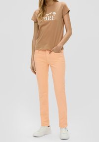 Maglietta di cotone marrone con grafica floreale bianca, abbinata a pantaloni di denim arancione chiaro. La modella indossa sneakers bianche, in piedi su uno sfondo neutro.