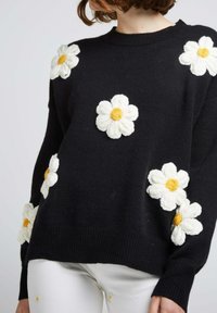Pull noir avec des appliqués de marguerites blanches et des centres jaunes, col rond et poignets côtelés. Le tissu semble doux et tricoté.