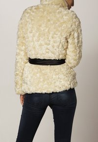 Sølvfarvet faux fur jakke med strukturerede, hævede mønstre og en sort bælte i taljen. Har en høj krave og lange ærmer.