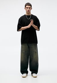 Cămașă neagră din bumbac supradimensionată, blugi largi, distresați, de culoare albastră, cu efect de decolorare, asortați cu adidași albi și un colier din argint.