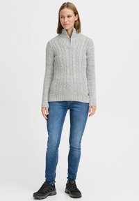 Grauer Kabelstrickpullover mit Stehkragen und Reißverschluss, kombiniert mit blauen Skinny-Jeans und schwarzen Sportschuhen. Glatte Textur und taillierte Passform.