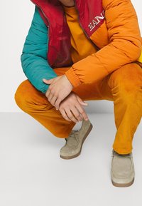 Chaqueta acolchada con secciones rojas y azules, pantalones de pana naranja y zapatos de ante beige, mostrando contrastes de color y texturas distintivos.