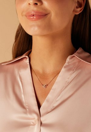 Goldene Kettenhalskette mit einem Schleifenanhänger, verziert mit kleinen Diamanten, getragen über einem rosa Satinhemd mit spitzem Kragen.