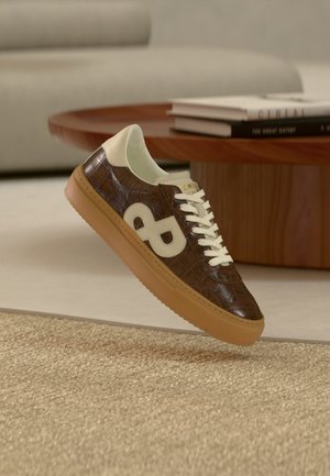 Zapatilla deportiva de cuero marrón con cordones crema y logo, suela de goma, colocada cerca de una mesa de madera con libros apilados en un salón beige.