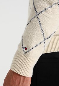 Maglione a trecce crema con motivi a rombi blu marino, polsini a costine e un piccolo logo sul maniche.