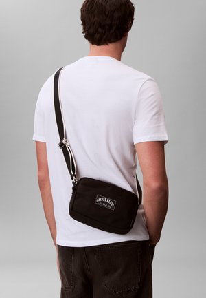 Άνδρας με λευκό t-shirt που φοράει μαύρη τσάντα ώμου με φαρδιά τιράντα και λογότυπο Calvin Klein στην πλάτη, σε απλό γκρι φόντο.