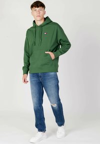 Felpa verde con tasca frontale e cordini, abbinata a jeans chiari strappati e sneakers bianche. Design semplice, vestibilità casual.