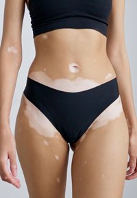Black bikini bottom with a low-rise cut, smooth texture, minimalistic design; worn with a matching black crop top. Skin shows vitiligo patches.