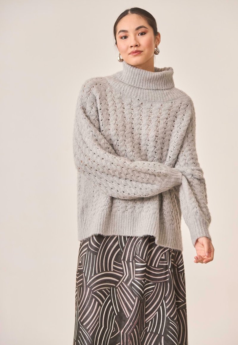 Suéter de punto gris con un alto cuello de cisne acanalado y patrones texturizados. Combinado con una falda estampada en tonos negros y plateados.