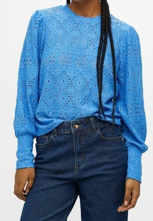 Femme portant un chemisier bleu vif en dentelle à manches longues avec des épaules bouffantes et un jean taille haute bleu foncé.