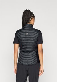 Černá zipová vesta s prošívaným vzorem na zádech, spárovaná s černým tričkem s krátkým rukávem. Obsahuje vysoký límec a hladkou texturu látky.