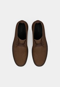 Rudos spalvos zomšiniai batai su lygiu paviršiumi, žemo kirpimo dizainu ir dviem dekoratyvinėmis raištelėmis priekyje.