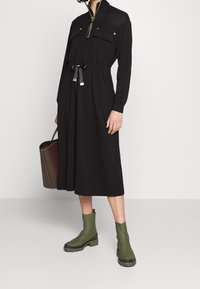 Black dress with a zip neck, two front pockets, and a cinched waist. Features long sleeves and a midi length. Paired with green ankle boots.