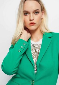 Groene blazer van gestructureerde stof, met een enkele knoopsluiting en drie decoratieve groene knopen op de manchetten, gecombineerd met een gemusterd top.