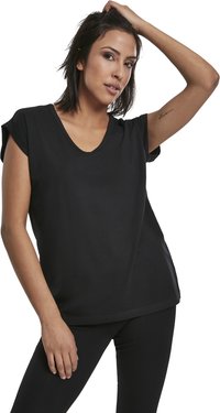 Una donna con capelli scuri di lunghezza media indossa una maglietta nera a maniche corte con scollatura a V e pantaloni neri, mentre posa con una mano sulla testa.