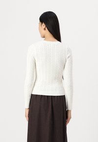 White knitted sweater featuring a cable pattern, crew neckline, and long sleeves, paired with a dark skirt.