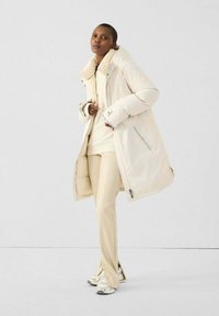 Mujer vestida con un abrigo acolchado largo color crema, pantalones a juego, sudadera con capucha y zapatillas blancas, de pie contra un fondo claro y sencillo.
